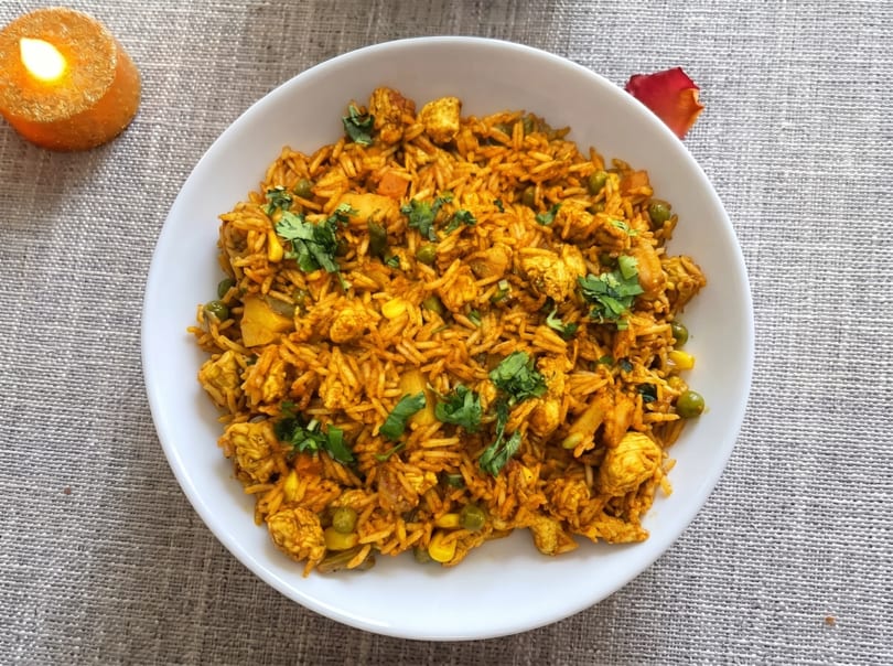 A white bowl of chicken biryani with peas, corn, and cilantro, next to a lit candle.