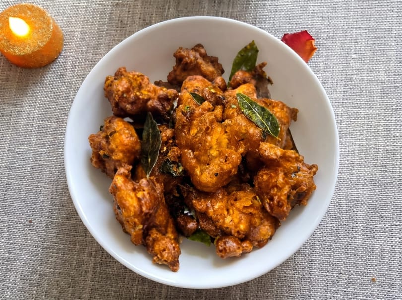 A white bowl filled with crispy, golden-brown fried chicken pakora garnished with curry leaves, next to a lit candle.