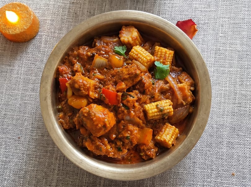 A brass bowl filled with a rich, orange-brown curry featuring baby corn, onions, and red peppers.