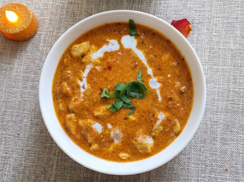 A white bowl of chicken curry with cream swirls and cilantro, next to a lit candle.